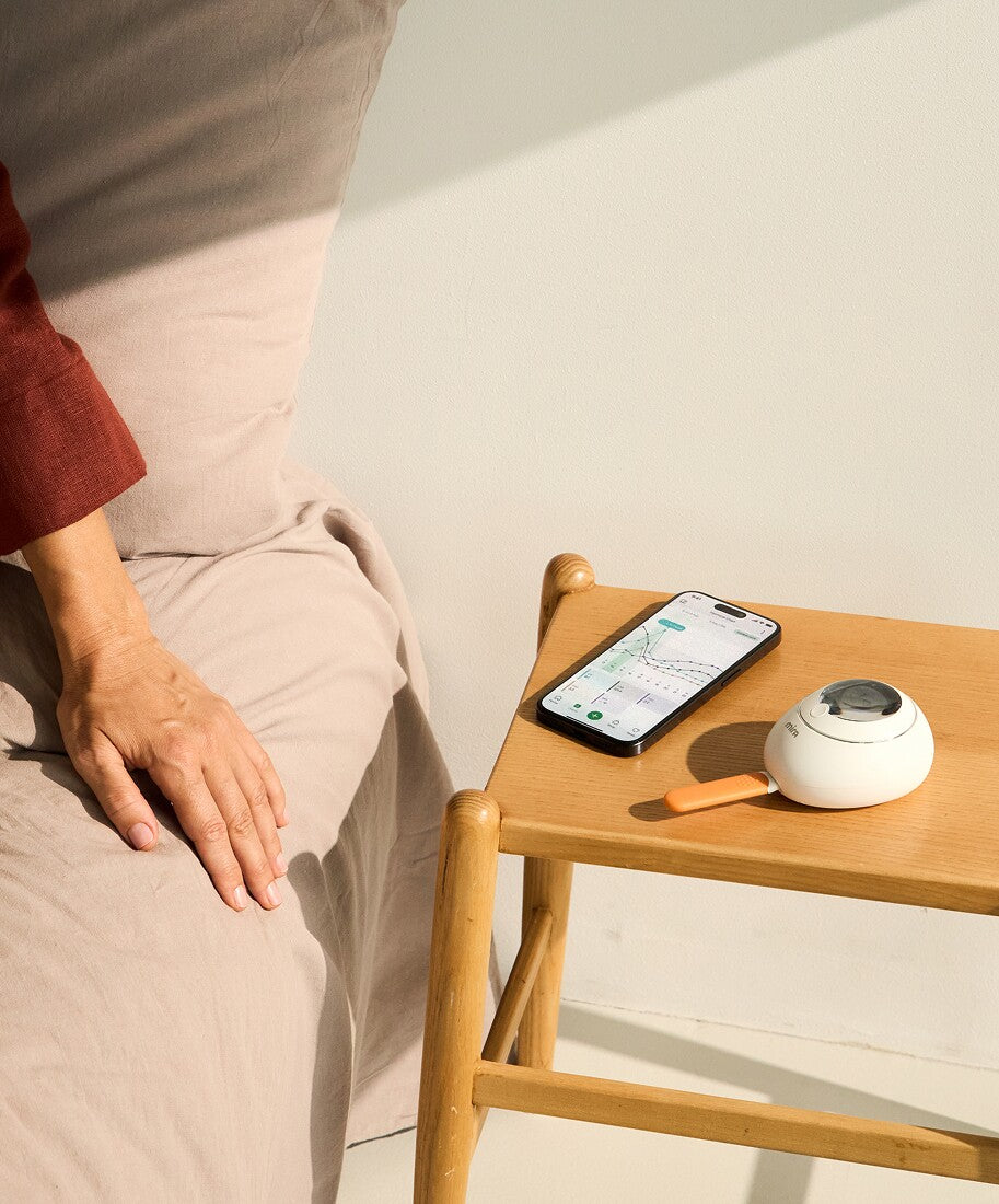 Mira Fertility Monitor and app on bedside table next to person resting in bed, tracking hormone data for fertility awareness