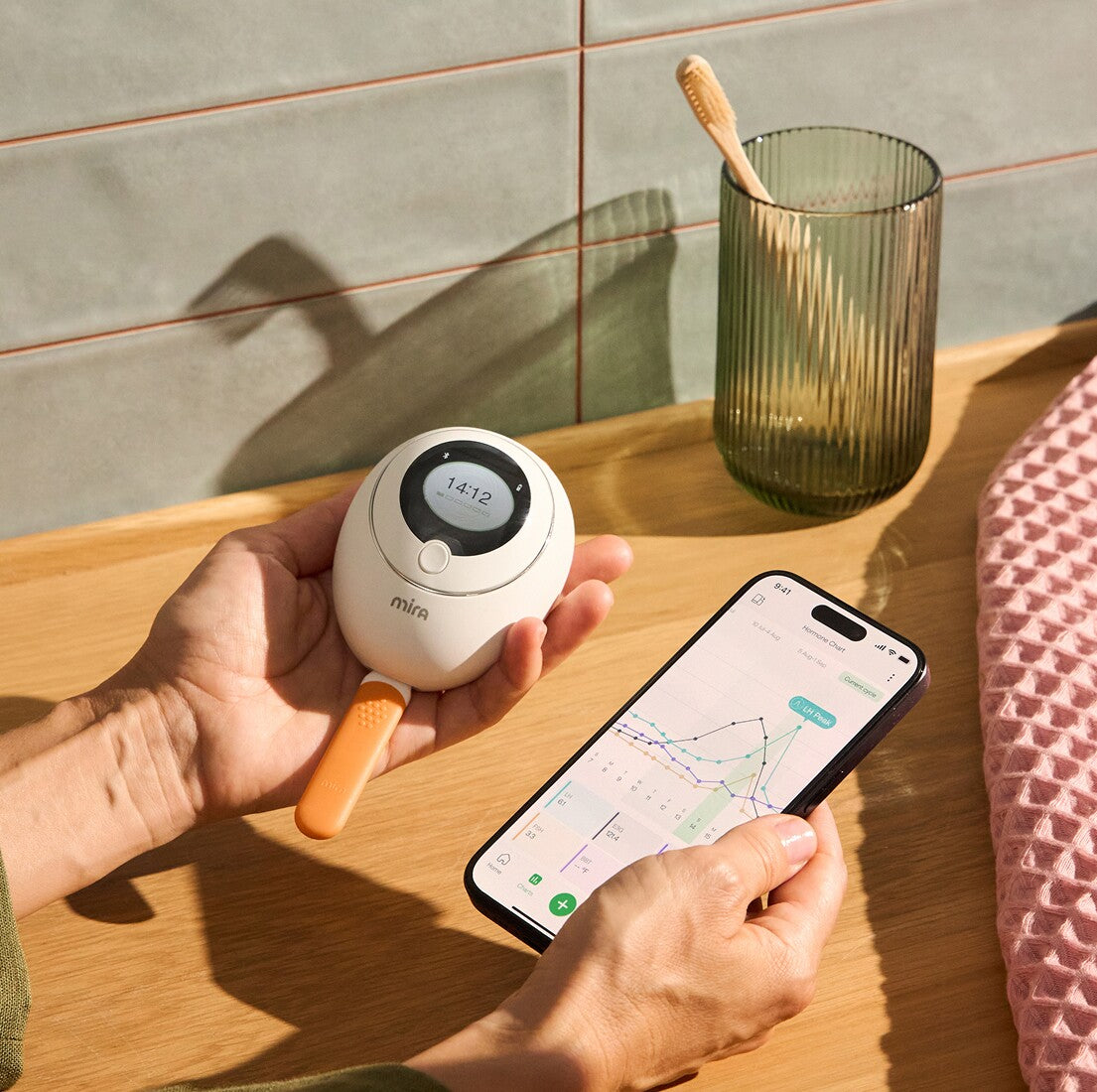 Hands holding Mira Fertility Monitor with wand and smartphone displaying hormone tracking chart with LH peak data