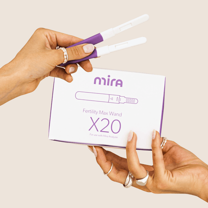 Hand holding two white fertility test wands with purple tips above Mira branded box labeled "Fertility Max Wand X20".