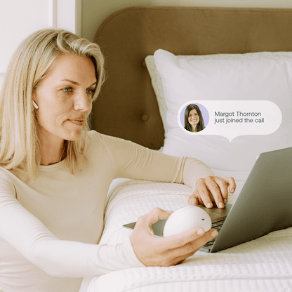 Woman using Mira fertility tracker device while on a telehealth video call from bed with laptop and AirPods