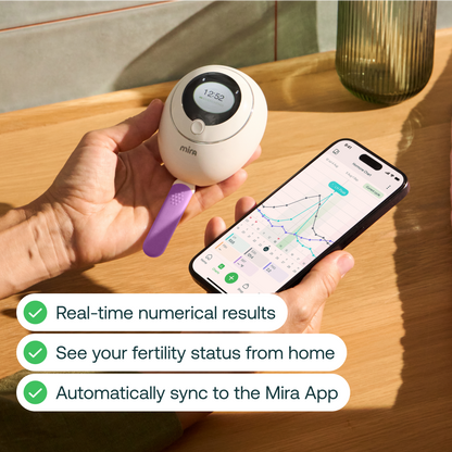 White handheld fertility test device with digital display held in hand next to smartphone showing app interface with graph.