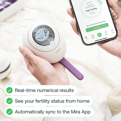 Hand holding white fertility testing device with purple handle next to smartphone displaying fertility tracking app.