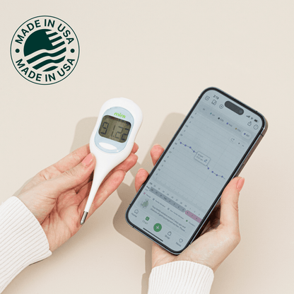 White Mira basal body thermometer with digital display held next to smartphone showing fertility tracking app.