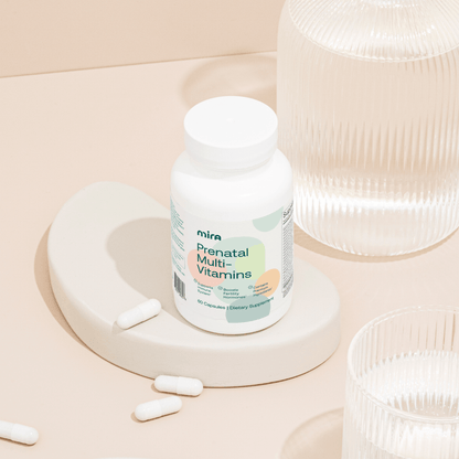 White bottle of Mira Prenatal Multi-Vitamins with capsules displayed on a cream-colored circular platform beside glass contai