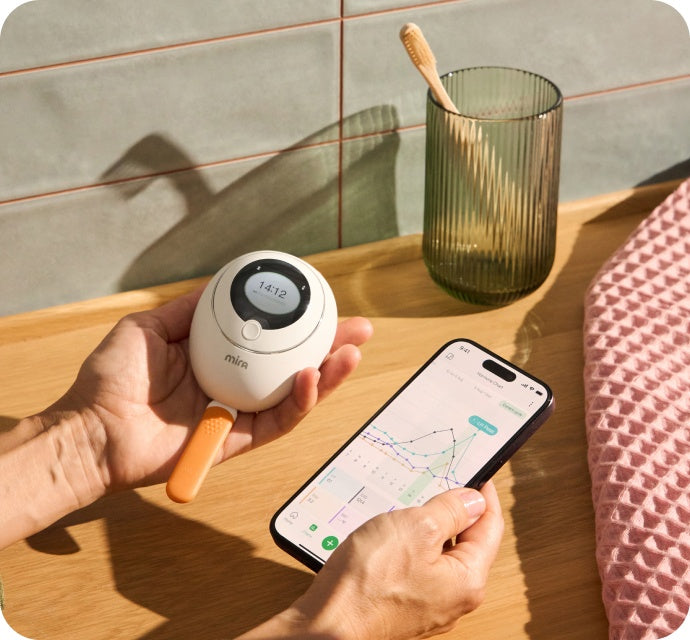 Hands holding Mira Fertility Monitor with wand and smartphone displaying hormone tracking chart with LH peak data