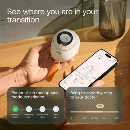 Hand holding white Mira hormone monitor device next to smartphone displaying hormone tracking data and graph on wooden table.