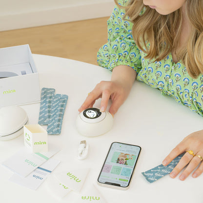 Child's hand touching white circular device next to smartphone, white box, blue patterned fabric, and documentation on table.