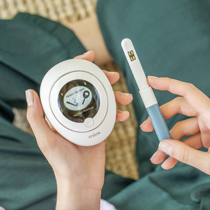 White circular Mira device with display screen and white pen-shaped applicator with blue grip held in hands.